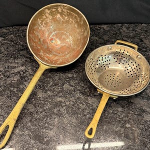 May include: A copper-colored bowl with a long, textured brass handle and a silver-toned metal colander featuring a perforated bowl, a short brass handle, and a small brass loop handle on the opposite side, both designed for kitchen use.