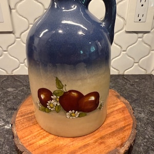 May include: A blue and cream-colored ceramic jug with a handle. The jug features a hand-painted design of three red plums with white daisies and green leaves. The jug is sitting on a round wooden slab.