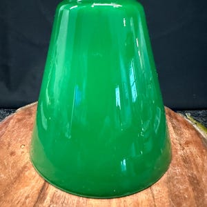 May include: A glossy, vibrant green glass lamp shade with a conical, tapered shape. The shade features a smooth, reflective surface and a circular opening at the top, designed to fit onto a light fixture base with a clean, minimalist profile.