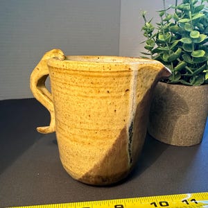 May include: A small, yellow ceramic pitcher with a unique, curved handle. The pitcher has a brown glaze on the bottom half and a yellow glaze on the top half. The pitcher is sitting on a black surface.