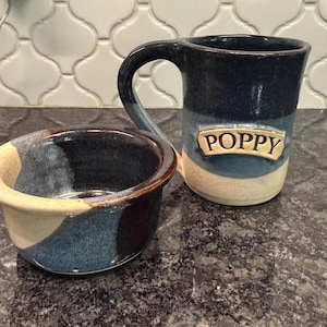 May include: A ceramic mug and a matching bowl featuring a tri-color glaze in dark blue, light blue, and cream. The mug has a large handle and a rectangular plaque on the front with the name POPPY embossed in capital letters in a serif font.