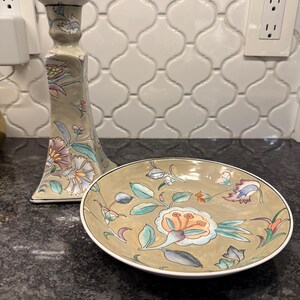 May include: A beige ceramic candle holder and a matching small plate. Both items feature floral designs in shades of pink, blue, and green. The candle holder is tall and slender, while the plate is round.