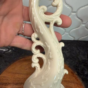 May include: Ivory-colored, ornate ceramic sculpture with a flowing, stylized design. The sculpture features swirling patterns and decorative flourishes. The piece has a glossy finish and appears to be a decorative item, possibly a candlestick or a similar ornamental object.