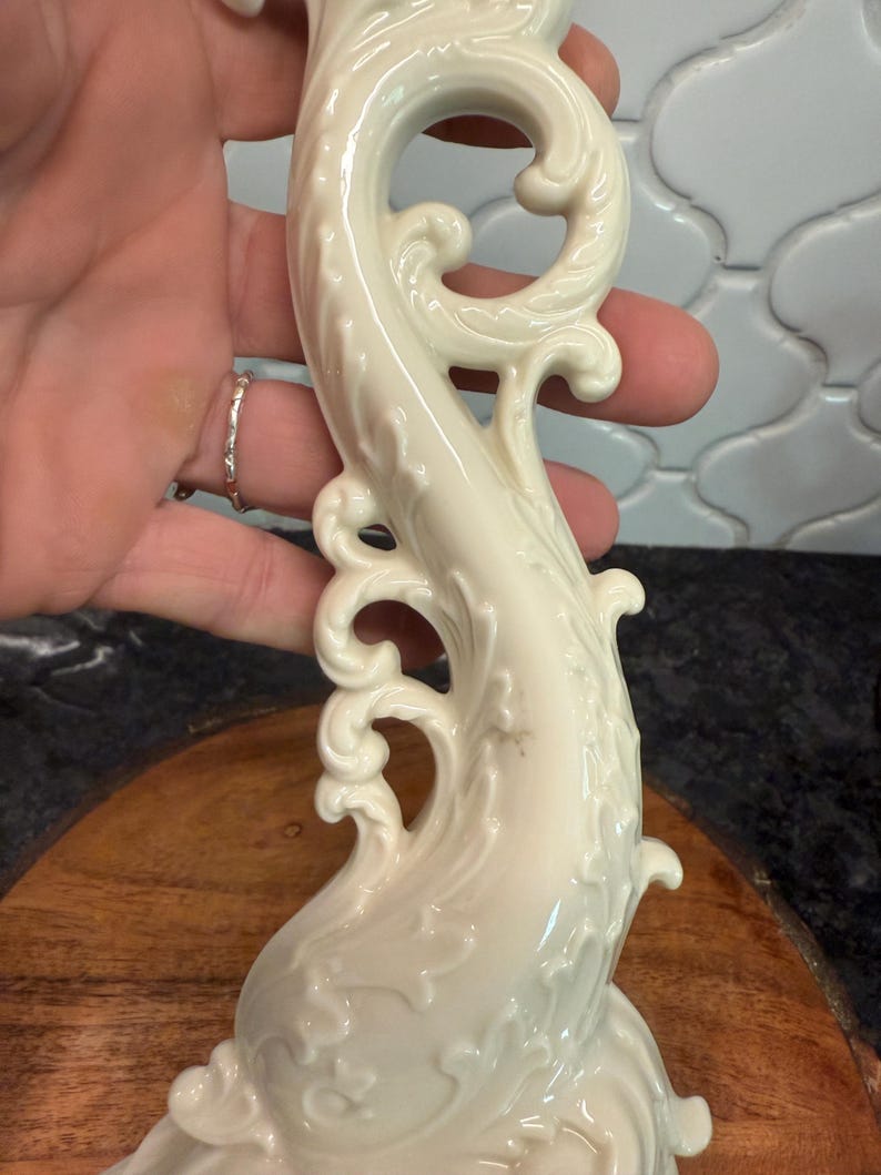 May include: Ivory-colored, ornate ceramic sculpture with a flowing, stylized design. The sculpture features swirling patterns and decorative flourishes. The piece has a glossy finish and appears to be a decorative item, possibly a candlestick or a similar ornamental object.