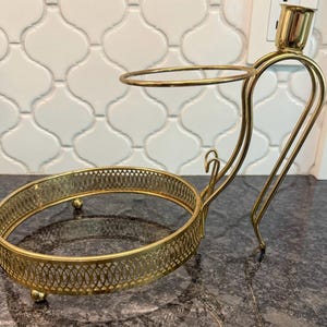 May include: A gold-colored, vintage-style candle holder with a circular base and a smaller ring above. The design includes a curved handle and a small cup for a candle. The base has a decorative, openwork pattern.