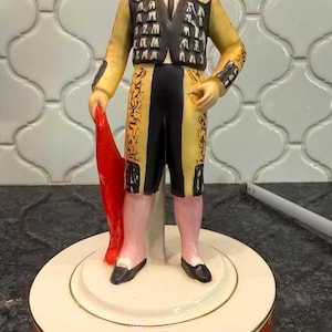 May include: A ceramic figurine of a matador in a traditional costume. The figure wears a black hat, a yellow jacket with black accents, and black shorts. The matador holds a red cape and stands on a round base with a red and gold pattern.
