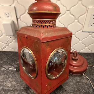 May include: A red metal lantern with a square body, a domed top, and circular glass windows on each side. The lantern features gold-toned trim around the windows and a decorative perforated band near the top. A separate matching red metal circular base piece sits nearby.