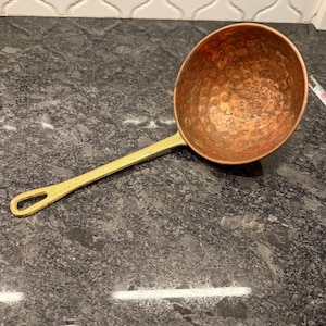 May include: A copper ladle with a hammered bowl and a brass handle. The handle has a loop at the end for hanging. The ladle is designed for kitchen use and is made of metal.