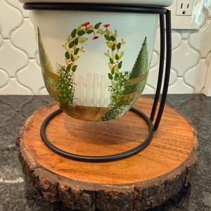 May include: A white and black metal planter with a garden scene painted on it, featuring a white picket fence and an archway of greenery. The planter sits on a round, wooden base with a black metal ring and a natural bark edge.