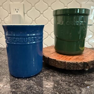 May include: Two Le Creuset stoneware utensil crocks, one in a vibrant blue and the other in a deep forest green. Both cylindrical containers feature a smooth, glossy finish, a flared rim, and the embossed Le Creuset logo on the front exterior.