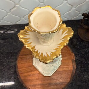 May include: A vintage, off-white vase with a gold-painted rim and leaf-like accents. The vase sits on a matching saucer with gold trim. The base is a light green color. The vase is displayed on a round, wooden surface.