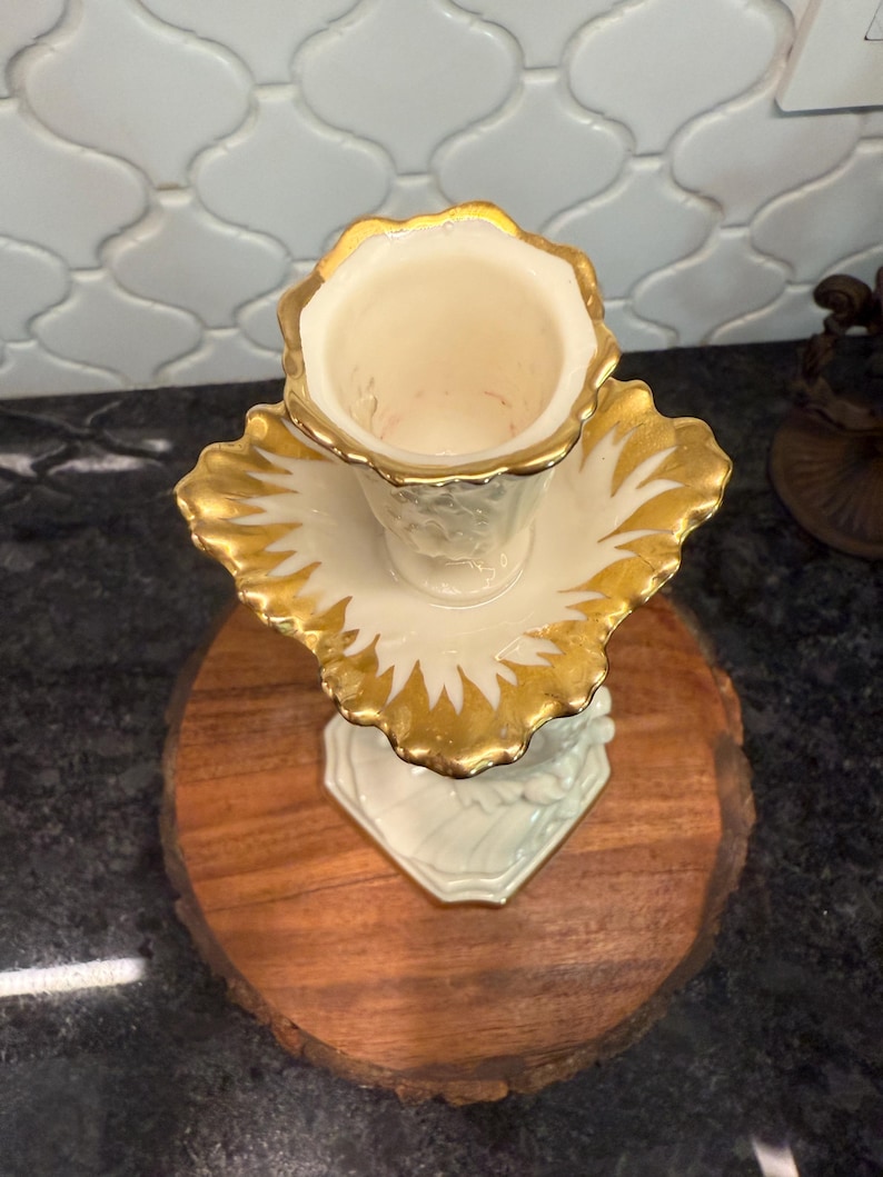 May include: A vintage, off-white vase with a gold-painted rim and leaf-like accents. The vase sits on a matching saucer with gold trim. The base is a light green color. The vase is displayed on a round, wooden surface.