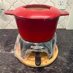 May include: A bright red enameled cast iron fondue pot with two side handles and a scalloped rim. The pot rests on a black metal stand labeled SWISSMAR, which is partially wrapped in clear plastic, all set upon a round light-colored wooden base.