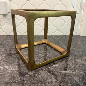 May include: A hollow, cube-shaped decorative frame with a metallic gold finish. The object features four vertical corner posts connected by horizontal bars at the top and bottom, creating an open, geometric structure with a textured, matte surface.