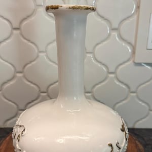 May include: A white ceramic decanter with a bulbous base and a long neck. The decanter features a decorative floral design in gold around the base and a white stopper. The decanter is displayed on a wooden slice.