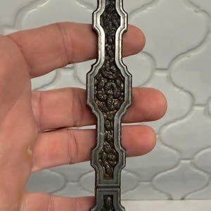 May include: A long, vertical metal cabinet pull with a dark, textured, hammered-style center inlay framed by a smooth, raised silver-toned border. The handle features a repeating geometric, elongated shape with rounded, scalloped edges.