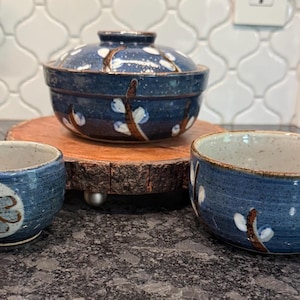 May include: A set of three blue ceramic bowls with a floral design. The set includes a lidded bowl and two smaller bowls. The bowls are decorated with white and brown floral accents and are set on a wooden base.