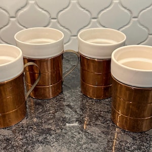 May include: Four copper-colored mugs with white ceramic tops and copper handles. The mugs have a cylindrical shape with horizontal bands. The mugs are sitting on a dark countertop.