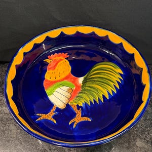 May include: A round ceramic bowl featuring a deep blue interior and a vibrant hand-painted rooster at the center. The rooster displays shades of red, orange, yellow, and green. The bowl has a scalloped rim painted in a bright yellow color.
