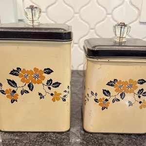 May include: Two vintage cream-colored metal canisters with black lids and floral designs. The canisters have clear glass knob handles. The floral design features orange flowers and black leaves. One canister is taller than the other.