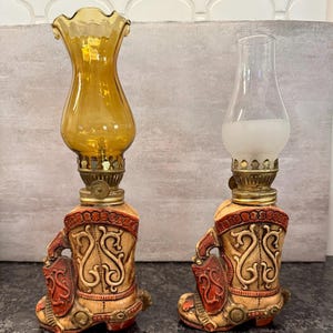 May include: Two decorative oil lamps. Each lamp features a cowboy boot base in beige, red, and brown tones. The lamps have a brass-colored metal base and a glass chimney. One lamp has a clear chimney, and the other has a yellow-tinted chimney.