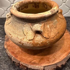 May include: A weathered, round, terracotta pot with two handles, resting on a circular wooden base. The pot has a wide opening and a textured surface, showcasing a rustic aesthetic. The wooden base has a natural bark edge.