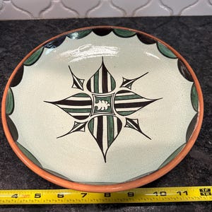 May include: A round ceramic plate with a light green center and a decorative black, green, and white starburst design. The rim is orange with green and black scalloped accents. The plate measures approximately 10 inches in diameter.