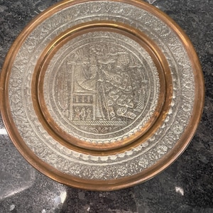 May include: A round, antique metal plate with an intricate engraved design. The plate features a silver-colored center with a detailed scene and a copper-colored rim. The design includes figures and floral patterns.