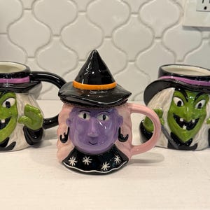 May include: Three ceramic mugs shaped like witch faces. Two mugs feature green-skinned witches with black hats and white hair. The center mug features a purple-skinned witch with a black hat, orange band, and pink hair, wearing a black collar with white stars.