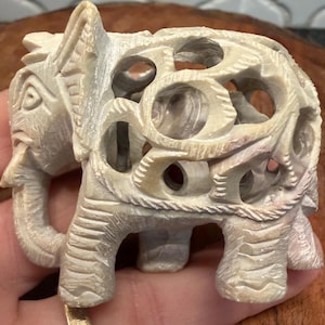May include: A carved stone elephant figurine in a light beige color. The elephant has intricate openwork detailing on its body and a trunk curled upwards. The figurine is standing on four legs and has a detailed head with visible eyes and ears.