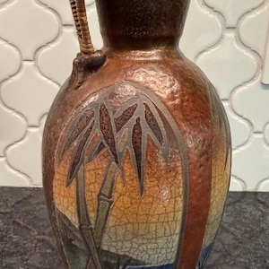 May include: A ceramic vase with a crackled glaze finish in shades of brown, orange, and yellow. The vase features a painted bamboo scene and a decorative handle wrapped in light brown wicker, with two small bamboo sticks tucked into the handle.