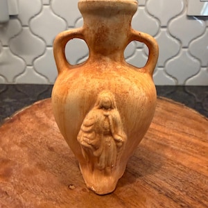 May include: A small, antique-style ceramic vase with a cork stopper. The vase is a light brown color with a textured surface and two handles. A carved figure is on the front of the vase. The vase is sitting on a wooden surface.