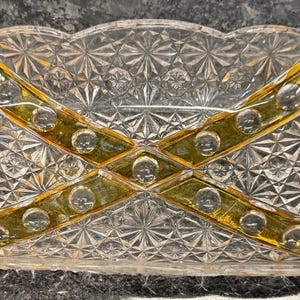 May include: A clear, rectangular glass dish with a scalloped edge and a diamond-cut pattern. The dish features gold-colored accents in an X-shaped design across the center. The dish is likely vintage and suitable for serving or display.