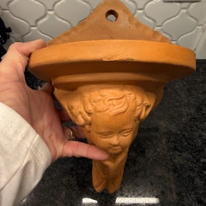 May include: A terracotta wall planter featuring a cherub face. The planter has a triangular top with a hole for hanging, a circular shelf, and a cherub face as the main decorative element. The color is a warm, earthy orange.