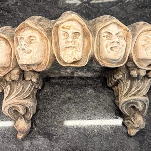 May include: A beige, ornate wall bracket featuring seven sculpted faces in hooded cloaks. The faces display various expressions. The bracket has a decorative, scrolling design on the supports and a dark gray background.