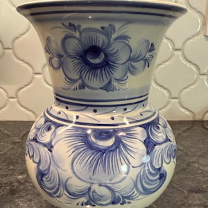May include: A ceramic vase with a bulbous base and flared rim, hand-painted with blue floral designs on a cream background. The vase features a detailed pattern of stylized flowers and leaves, suitable for home decor.