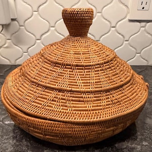May include: A woven rattan basket with a conical lid and a handle. The basket is a warm, natural brown color and has a textured surface. The basket is likely used for storage or decoration.