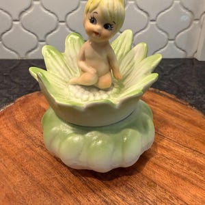 May include: A ceramic figurine of a seated fairy with blonde hair and blue eyes, atop a light green flower-shaped container. The container sits on a larger, matching green base, all on a wooden surface. The figurine is approximately 3 inches tall.