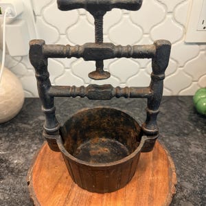 May include: An antique, cast iron fruit press with a wooden base. The press features a screw mechanism and a small, round bowl. The metal has a rustic, aged appearance, with visible signs of wear and rust. The wooden base is a natural, circular slice.