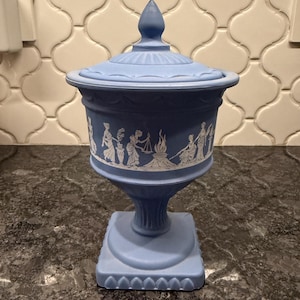 May include: A light blue lidded urn with a square base and a pedestal. The urn features a white relief design of classical figures and a central flame. The lid is topped with a pointed finial. The urn is likely made of ceramic or porcelain.