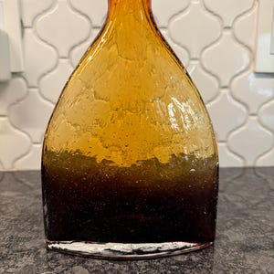 May include: Amber-colored glass vase with a textured surface. The vase has a wide, flat base that tapers upwards to a narrow neck. The color transitions from a dark brown at the base to a golden amber at the top. The vase is likely used for decorative purposes.