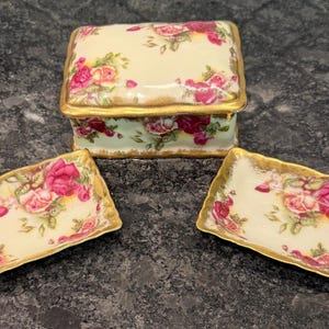 May include: A decorative porcelain box with a lid and two matching rectangular dishes. The set features a cream-colored background with pink and red rose floral patterns and gold trim. The box is rectangular with a gold trim.