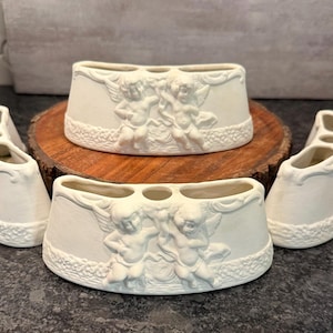 May include: A collection of off-white ceramic toothbrush holders. Each holder features a detailed relief of cherubs and a decorative border. The holders are arranged on a wooden surface and a dark countertop.