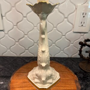 May include: A cream-colored ceramic candlestick holder with gold trim. The candlestick has a detailed floral design and is placed on a wooden round. The candlestick is designed to hold a single candle.
