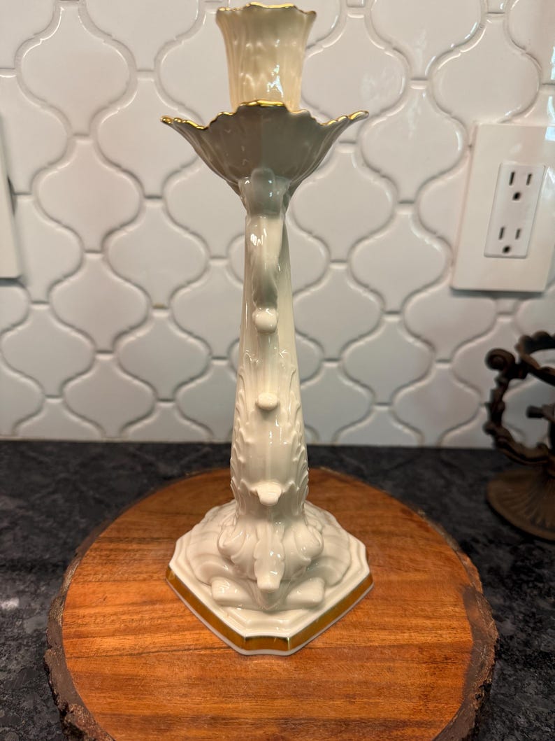May include: A cream-colored ceramic candlestick holder with gold trim. The candlestick has a detailed floral design and is placed on a wooden round. The candlestick is designed to hold a single candle.