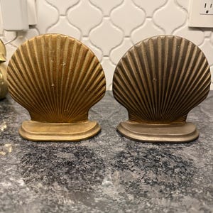 May include: Two seashell-shaped bookends. One is gold-colored, the other is bronze. Both have a fan-like design with vertical lines and a flat base. They are displayed on a dark, speckled surface.