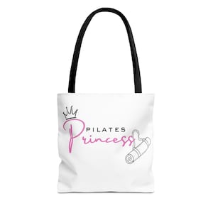 May include: White tote bag with black handles featuring the text "PILATES Princess" in pink script, a crown, and a line drawing of a rolled-up yoga mat. The bag is designed for carrying items.