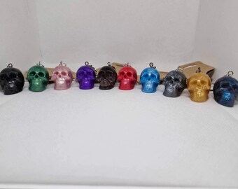 May include: A collection of nine skull-shaped pendants in various colors, including black, green, pink, purple, brown, red, blue, gray, and gold. Each skull has a small metal loop at the top for hanging. The pendants are arranged in a row.