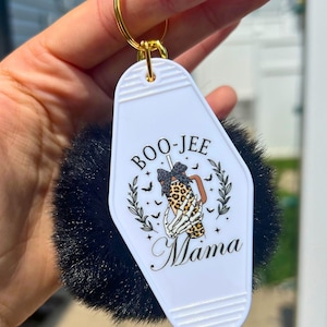 May include: White motel key chain with a black and white skeleton hand holding a coffee cup with a leopard print design. The text on the keychain says "Boo-Jee Mama".