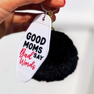 May include: White motel key chain with black text that reads "Good Moms Say Bad Words" and a black fluffy pom-pom attached.